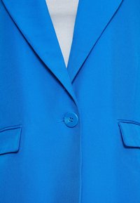 Blazer bleu vif avec un seul bouton bleu fermé, poches à rabat, et un t-shirt blanc côtelé visible en dessous.