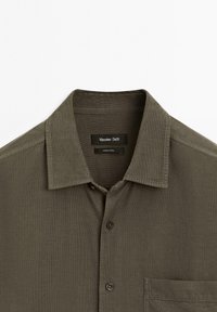 Olivově zelená košile s límečkem na knoflíky, krátkými rukávy, jednou náprsní kapsou a texturovanou látkou. Štítek nese nápis "Massimo Dutti."