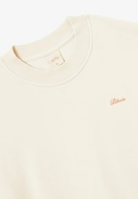 Crème sweatshirt met geribbelde ronde hals, voorzien van een klein geborduurd merklogo in goud aan de voorkant. Zachte textuur, relaxed ontwerp.