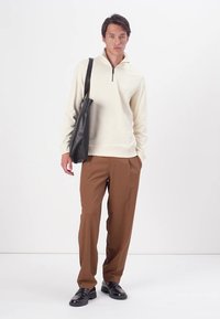 Light beige sweatshirt with a half-zip collar, brown trousers, and black shoes; model holds a black leather bag. Simple, relaxed fit.