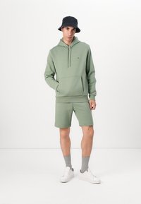 Groene katoenen hoodie met een voorzak en een trekkoord, gecombineerd met bijpassende groene shorts. Gemaakt met een zwarte vissershoed en witte sneakers.