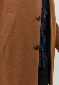 Cappotto di lana marrone con una texture morbida, dotato di una tasca laterale e bottoni neri, indossato sopra una camicia di jeans blu navy.