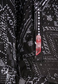 Gros plan sur un tissu noir avec des motifs géométriques blancs complexes et une étiquette verticale rouge et noire portant l'inscription "TOK".