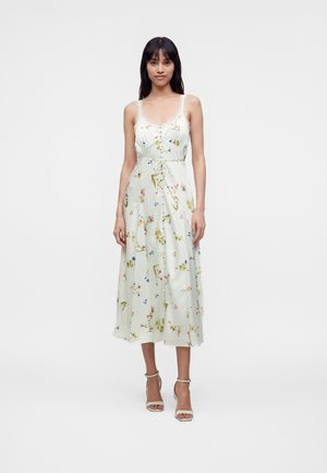 Femme portant une robe midi blanche sans manches avec imprimé floral, bordure en dentelle, boutons sur le devant et taille nouée, associée à des sandales beiges à talons hauts.