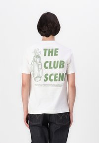 Hvid kortærmet t-shirt med et grønt grafisk motiv af en golftaske og teksten "THE CLUB SCENE" på ryggen. Løs pasform, blødt bomulds materiale.