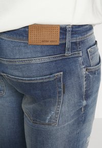 Jeans de denim azul con un parche perforado beige que lleva la marca "ANTONY MORATO" en la cintura y múltiples bolsillos en la parte posterior.