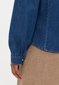 Denimskjorte med lange, puffete ermer; mørkeblå farge, knappemansjettdetalj og buet nederkant, båret over et teksturert, lyst brunt stoff.