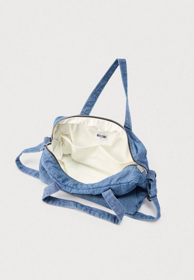 Modra denim torbica za čez ramo z nastavljivim trakom, odprt zadrga razkriva notranjost iz kremaste tkanine in etiketo Moschino Baby na notranji strani.