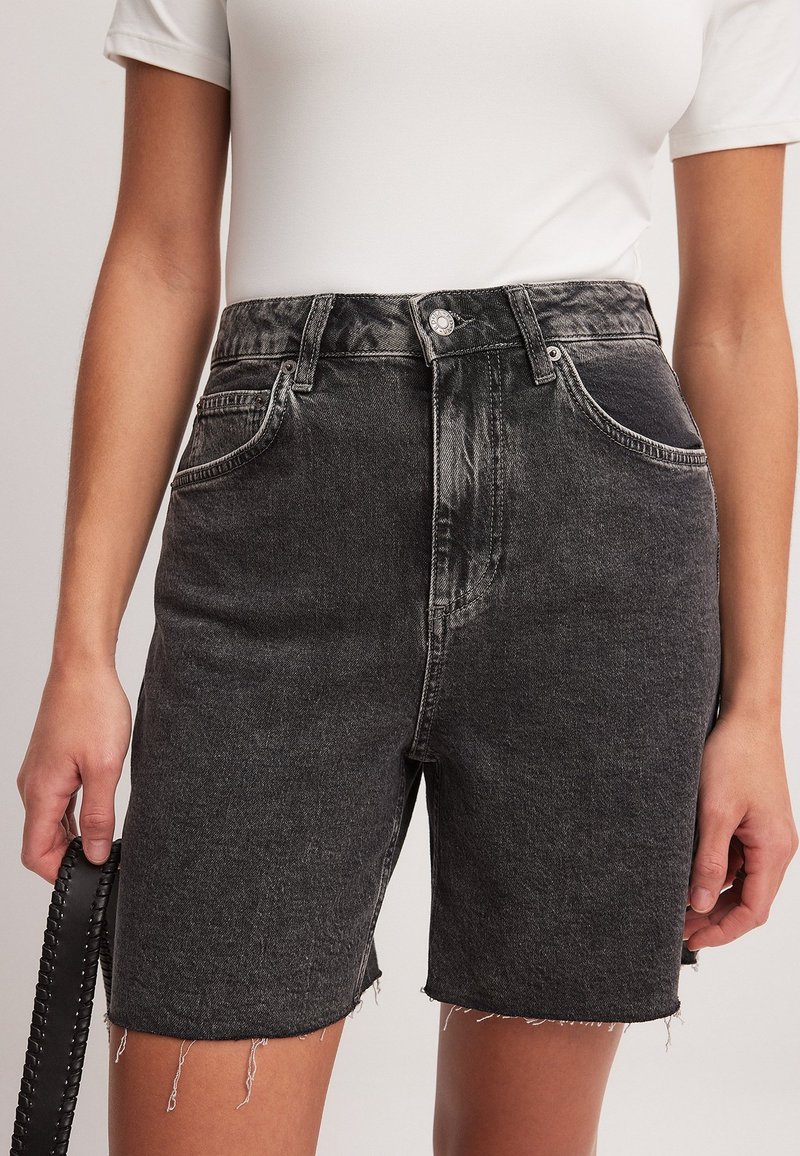 Moteris vilki juodus džinsinius šortus su dideliu liemeniu ir pravirais kraštais bei baltą prigludusią trumpomis rankovėmis palaidinę, laikydama juodą pintą diržą.