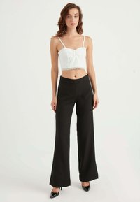 Hvid cropped top med blondekant og justerbare stropper, parret med sorte vide bukser. Modellen bærer sorte højhælede sko.