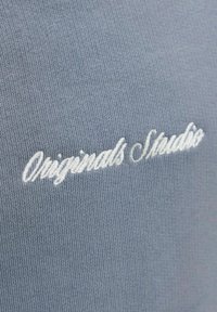 Ελαφρύ γαλάζιο ύφασμα με λευκό κεντημένο κειμενό σε γραφή με πλάγια γράμματα που γράφει "Originals Studio."