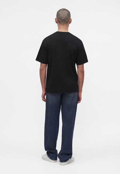 Homme aux cheveux courts portant un t-shirt noir uni, un jean bleu foncé ample et des baskets blanches, debout de dos sur un fond uni.