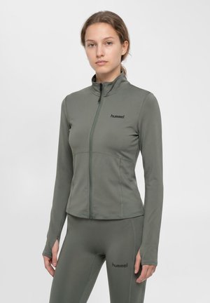 Młoda kobieta ubrana w dopasowaną oliwkowozieloną kurtkę zapinaną na zamek i legginsy z logo hummel, stoi na tle jednolitego jasnego tła.