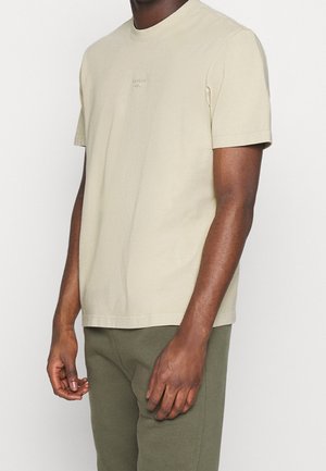 Mand iført en beige kortærmet Reebok t-shirt og olivengrønne bukser, stående foran en ensfarvet hvid baggrund.