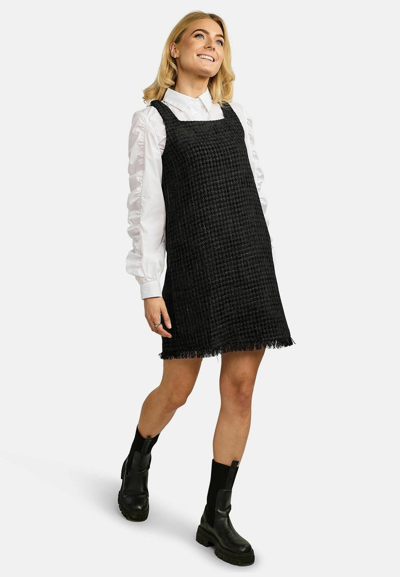 Black woven dress with a square neckline and fringe hem, paired with a white ruffled shirt and chunky black boots.