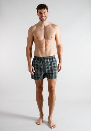 Boxer shorts in dark green plaid pattern with black accents, elastic waistband, and a single button. Model stands barefoot against a plain background.