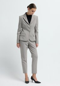 Femme portant un blazer ajusté gris clair et un pantalon assorti, un top noir, des escarpins noirs texturés, regardant vers le bas contre un fond clair uni.