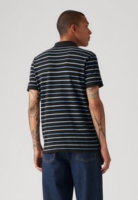 Black and blue striped polo shirt with a collar, short sleeves, and a relaxed fit. Cotton material with visible tattoos on the arms.