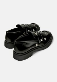Zwarte instapschoenen met decoratieve metalen knopen, met een gladde, glanzende afwerking en een dikke rubberen zool, in een schuine positie ter presentatie.