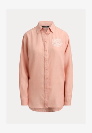 Koszula w kolorze brzoskwiniowym, z długim rękawem i zapięciem na guziki, z kołnierzykiem na spiczaste końce oraz białą, haftowaną inicjałą na lewej piersi.