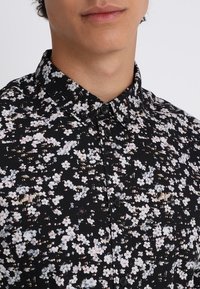 Musta kukkakuvioinen paita, jossa on napillinen kaulus, valkoisia ja pastellinsävyisiä kukkakuoseja sekä sileä ja kevyt kangaspinta.