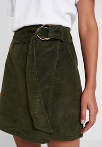 Mujer con pantalones cortos de pana verde oliva de cintura alta, con un cinturón ancho y hebilla dorada, combinados con una camiseta blanca metida por dentro.