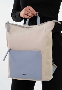 Bolsa con cuerpo crema, bolsillo frontal azul claro y asas grises; cuenta con un cierre de cremallera y el logo "TMRS" en el bolsillo.