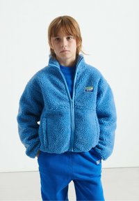 Chaqueta de forro polar azul con cremallera en el frente, dos bolsillos frontales y tejido texturizado. Combinada con una sudadera azul y pantalones. Presenta una etiqueta de la marca.