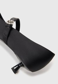 Zapato de tacón alto negro con un exterior de satén elegante, punta afilada, tira delgada en el tobillo y detalle metálico cerca de la hebilla.