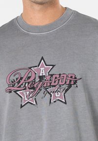 Šedé tričko s krátkým rukávem, které má grafický design s růžovými a černými hvězdami, stylizovaný text "Legacy" a "GmbH" v opotřebovaném stylu.