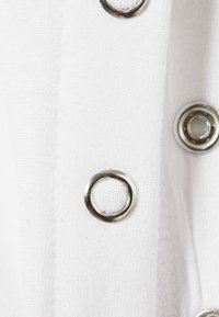 Λευκό ύφασμα με μεταλλικά ρούμπια. Η υφή είναι λεία με ορατές γραμμές ραφής. Τα μεταλλικά στοιχεία είναι κυκλικά και λαμπερά.