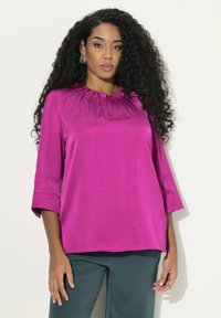 Blusa di satin fucsia con maniche a tre quarti, scollo arricciato e vestibilità ampia, abbinata a pantaloni verde scuro.