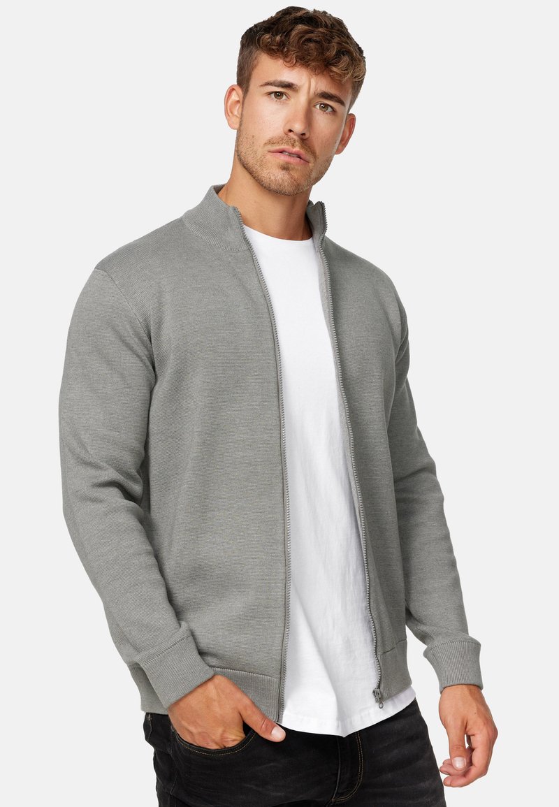 Grauer Zip-Up-Cardigan mit hohem Kragen, gerippten Bündchen und Saum. Trägt man über einem weißen Hemd. Der Stoff wirkt glatt und leicht.