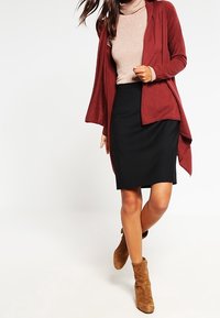 Cardigan rouge sur un col roulé beige, associé à une jupe crayon noire et des bottines chelsea en daim beige. Tricot texturé et silhouette ajustée.
