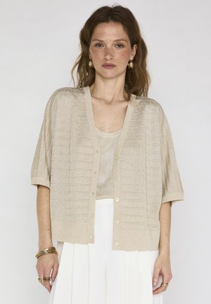 Femme aux cheveux bruns ondulés portant des boucles d'oreilles créoles en or, un cardigan beige à manches courtes boutonné, un haut assorti, une jupe blanche, ainsi que des bracelets et des bagues en or.