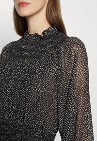 Femme portant un chemisier noir transparent à pois blancs, avec un col froncé à volants et des manches longues froncées.