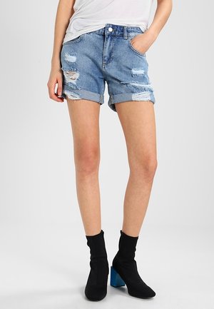 Person trägt hochgekrempelte, zerrissene Jeansshorts, weißes Oberteil und schwarze Stiefeletten mit blauen Blockabsätzen, steht mit einer Hand in der Tasche.