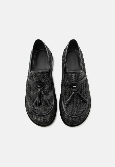 Ζευγάρι μαύρα πλεκτά δερμάτινα μοκασίνια με φουντίτσες, θέα από πάνω σε λευκό φόντο.