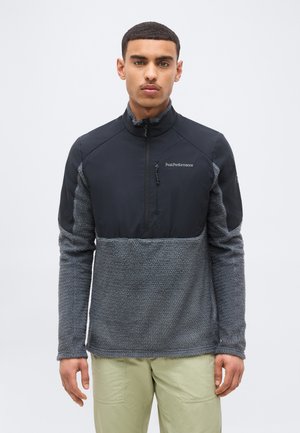 Vyras dėvi juodą ir pilką fliso striukę su priekiu užtrauktuku ir smėlio spalvos kelnes, stovintis priešais paprastą šviesų foną.