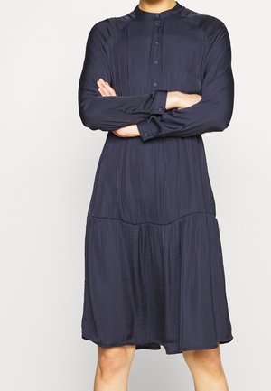 Personne aux bras croisés portant une robe bleu marine à manches longues, boutonnée, longueur genou, avec une partie jupe froncée.