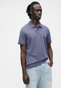 Polo de manga curta para homem em algodão azul escuro com corte ajustado, apresentando um pequeno logótipo amarelo bordado no peito. Jeans claros.