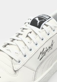 Hvite lær sneakers med grå detaljer, med flate hvite snøringer, runde snørehull og et logomerket på tungen. Teksten "Majesty" er brodert.
