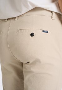 Beige bukser har et tekstureret stof, en baglomme med en sort knap og et lille mærkat nær lommen.