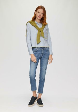 Gestreiftes blaues Hemd mit Knöpfen, grüner Strickpullover um die Schultern gebunden, ausgefranste hellblaue Jeans und schwarze Sneakers mit dicker Sohle.