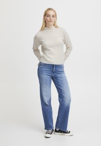 Maglione lavorato a maglia beige chiaro con colletto alto, abbinato a jeans a gamba dritta blu chiaro e sneaker nere. Outfit semplice e casual.