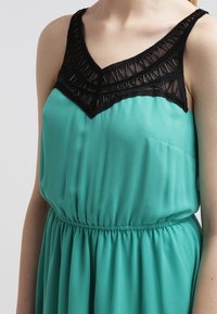 Tyrkysové šaty bez rukávů se sbíraným pasem. Černá krajková tělová část s geometrickými vzory a výstřihem do V. Hladká textilie.