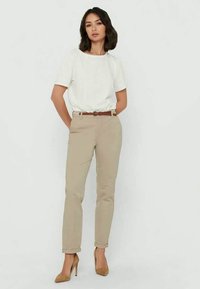 Beige trousers with a slim fit and a brown belt. The model wears a white, short-sleeved shirt and tan heels, standing upright against a neutral background.