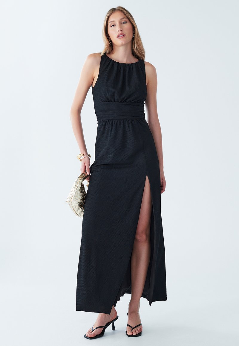 Schwarzes Maxikleid mit rundem Ausschnitt, gerafftem Oberteil und einem Seitenschlitz. Getragen mit schwarzen Riemchensandalen und einer geflochtenen Tasche.