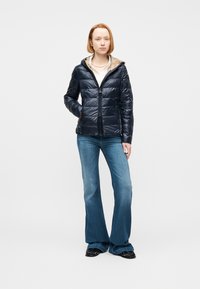 Νεαρή γυναίκα που φοράει μπλε μπουφάν puffer με μπεζ κουκούλα, λευκό πουκάμισο, μαργαριταρένιο κολιέ, μπλε καμπάνες τζιν και μαύρα παπούτσια, στέκεται με το ένα χέρι στην τσέπη.