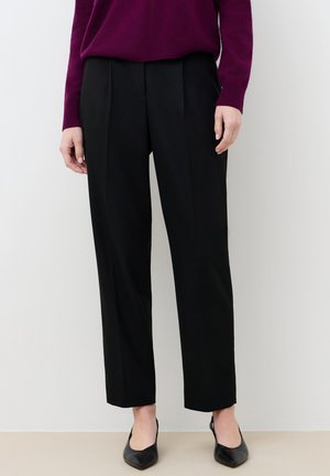 Osoba ubrana w czarne dopasowane spodnie, czarne buty w szpic oraz długi fioletowy sweter, stojąca na tle gładkiej ściany.